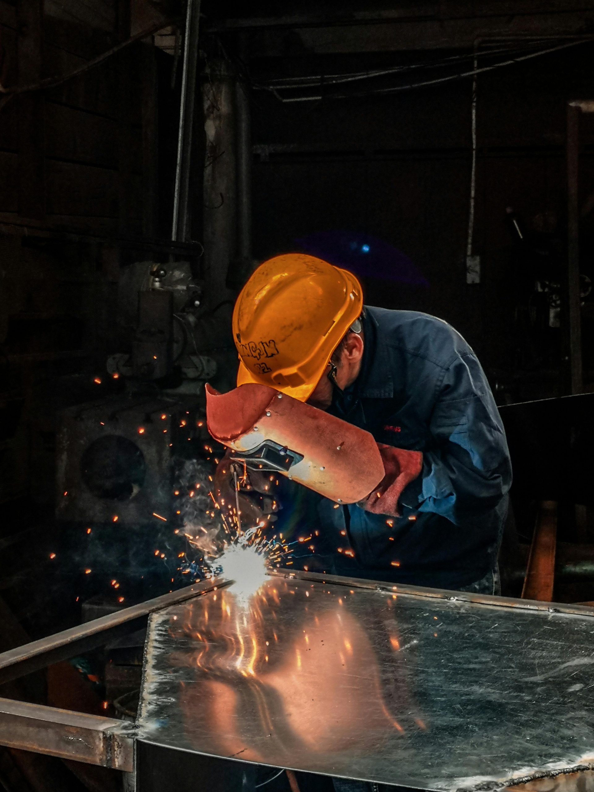 A welder wearing protective gear works on metal in an industrial workshop, surrounded by sparks.